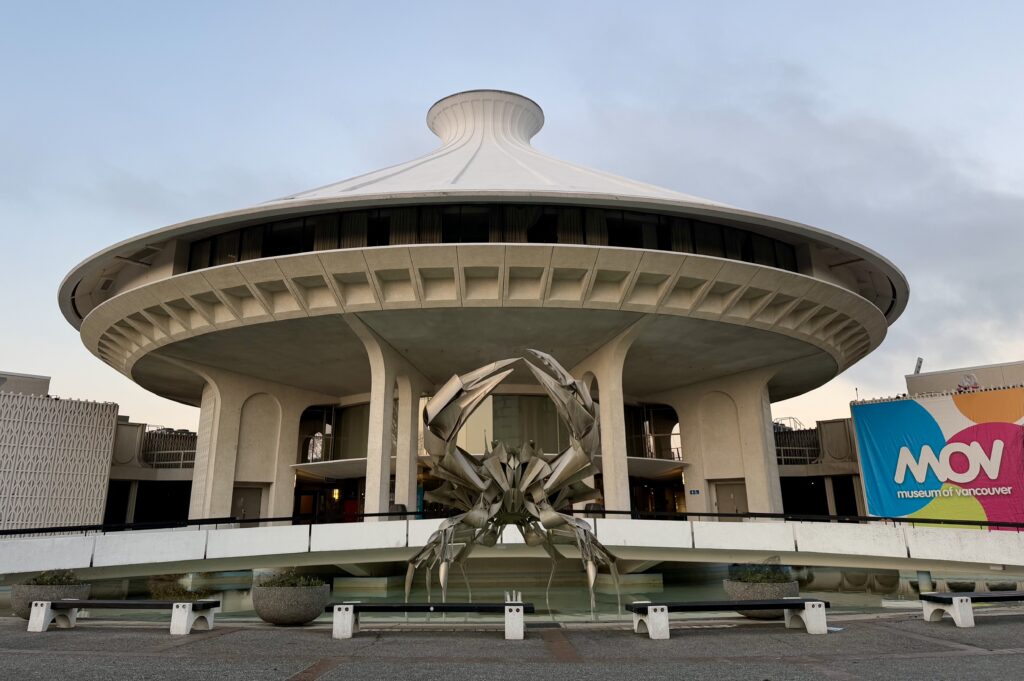  Museum of Vancouver, a distinctive round building with a futuristic, flying-saucer-like design. In the foreground, a striking metallic sculpture of a geometric crab stands in a reflective water feature, adding an artistic and dramatic element. To the right, a colorful banner with the museum's name, "MOV Museum of Vancouver," is displayed, making the location clear. The composition captures the building’s bold architecture and the interplay of light and shadow at what appears to be early morning or late afternoon. 